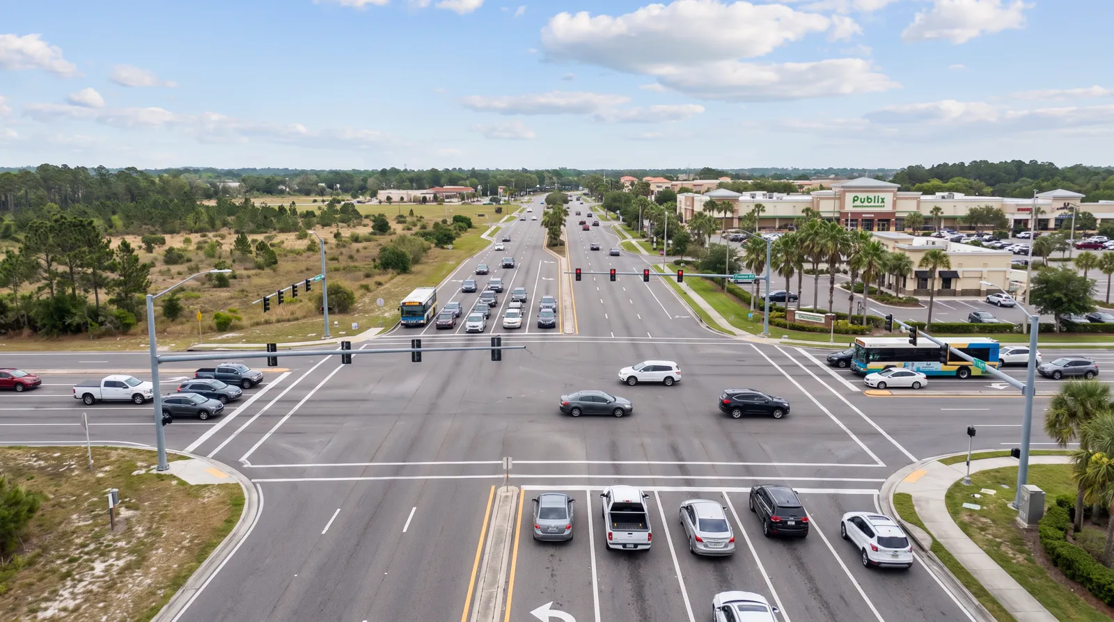 A wide daytime view of the Cortez Boulevard and Barclay Avenue intersection in Spring Hill, showing six lanes of traffic beside the Publix-anchored shopping center and open land under a bright Florida sky.